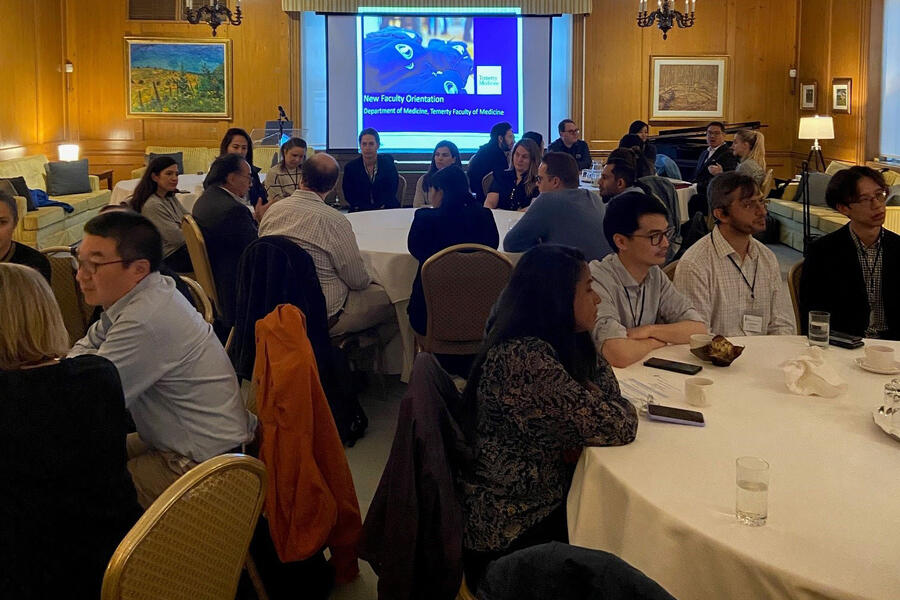 Group of people sitting at tables in a room with a screen displaying "New Faculty Orientation" in the background