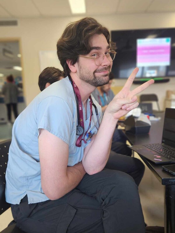 white man with brown hair, wearing blue scrubs and giving a peace sign