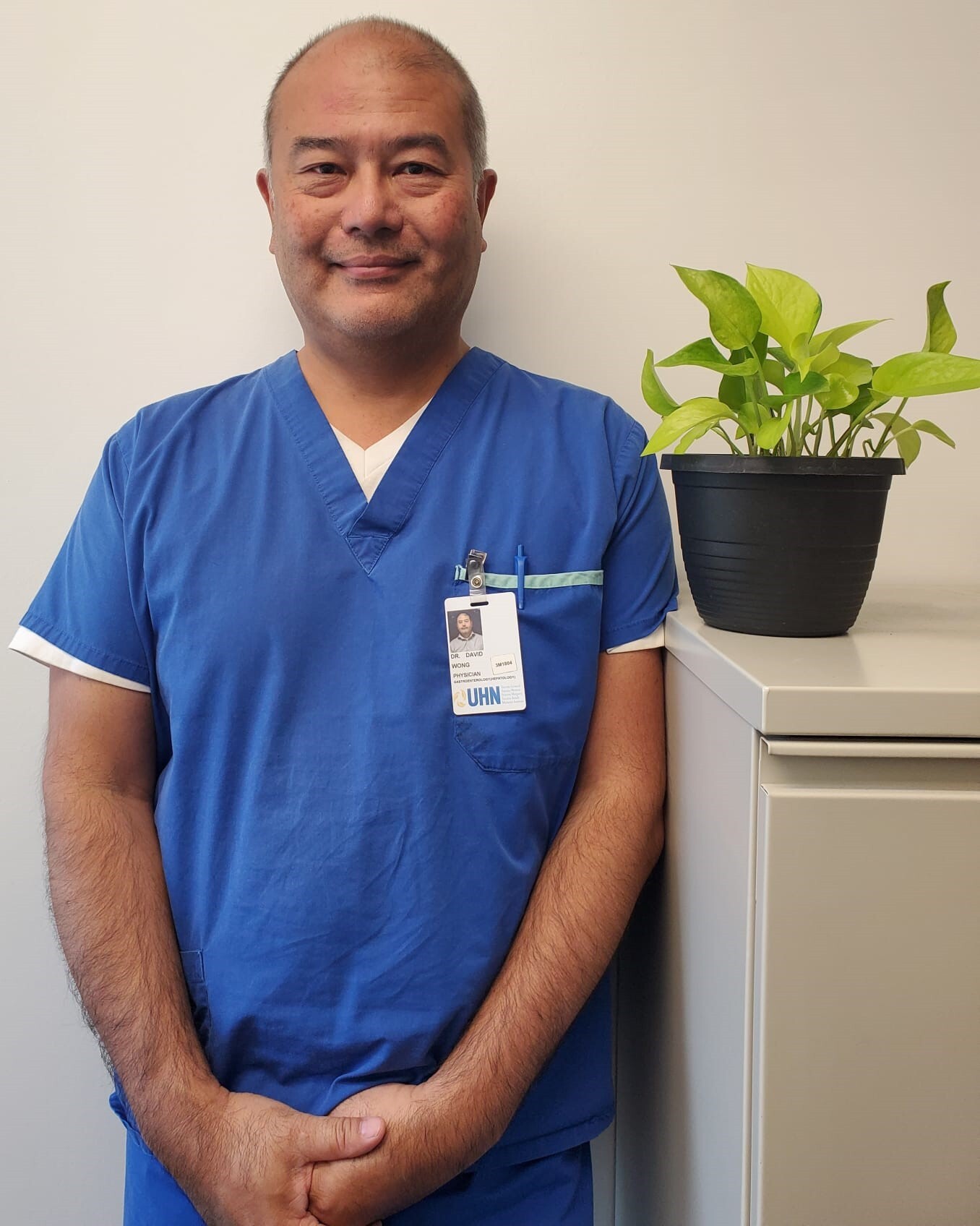 A smiling Dr. David Wong wearing blue scrubs while standing next to a leafy plant on a filing cabinet.