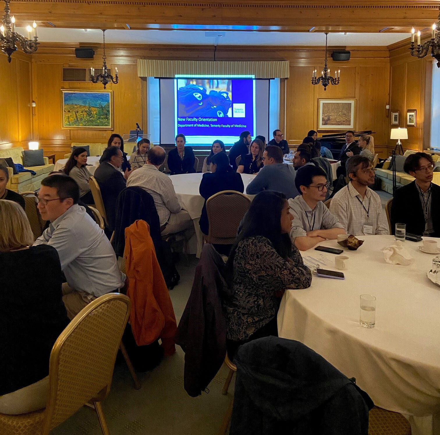 Group of people sitting at tables in a room with a screen displaying "New Faculty Orientation" in the background